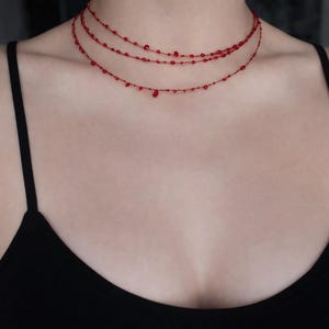 Peut inclure: Un collier à plusieurs rangs avec trois rangées de petites perles rouges. Le bijou délicat est porté sur un haut noir, créant un contraste. Les perles sont espacées uniformément, avec quelques perles plus grosses ajoutant des détails.