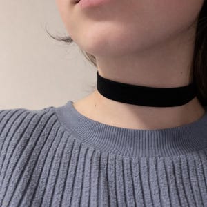 May include: A black velvet choker necklace. The choker is worn around the neck and is a simple, elegant accessory. The person is wearing a blue ribbed sweater.