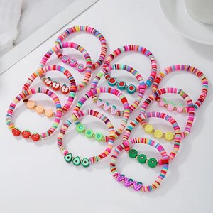 May include: A collection of colorful beaded bracelets. Each bracelet features a rainbow striped design and is adorned with fruit-shaped charms, including strawberries, lemons, and avocados. The bracelets are displayed on a white surface.