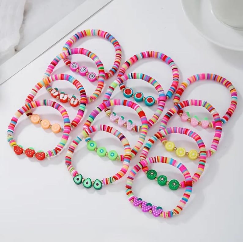May include: A collection of colorful beaded bracelets. Each bracelet features a rainbow striped design and is adorned with fruit-shaped charms, including strawberries, lemons, and avocados. The bracelets are displayed on a white surface.