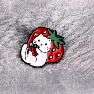 May include: A cute enamel pin featuring a white cat holding a strawberry, set against a red strawberry with green leaves. The pin has a black outline and is set on a gray background.