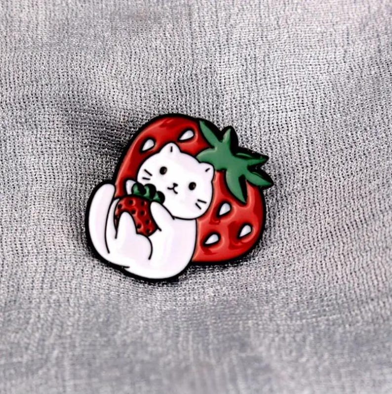 May include: A cute enamel pin featuring a white cat holding a strawberry, set against a red strawberry with green leaves. The pin has a black outline and is set on a gray background.