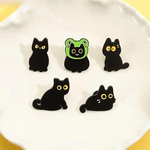 May include: A collection of five black cat-themed pins. The pins feature various cat poses, including one wearing a green frog hat. Each cat has yellow eyes. The pins are on a white, scalloped plate.