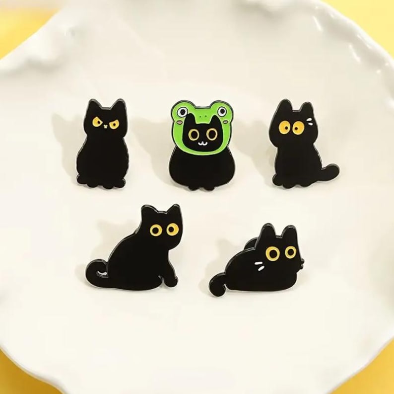 May include: A collection of five black cat-themed pins. The pins feature various cat poses, including one wearing a green frog hat. Each cat has yellow eyes. The pins are on a white, scalloped plate.