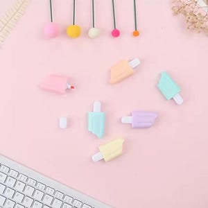 May include: A collection of pastel-colored popsicle-shaped highlighters and a pink highlighter pen on a light pink surface. The highlighters are in shades of pink, yellow, blue, and purple, with white popsicle sticks. A white keyboard is visible in the lower left corner.