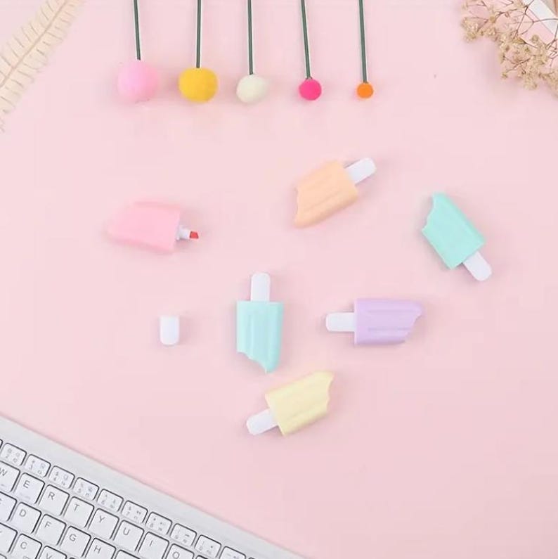 May include: A collection of pastel-colored popsicle-shaped highlighters and a pink highlighter pen on a light pink surface. The highlighters are in shades of pink, yellow, blue, and purple, with white popsicle sticks. A white keyboard is visible in the lower left corner.