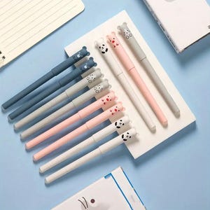 May include: A collection of colorful pens with animal-themed toppers. The pens are arranged on a white surface against a light blue background. The pens feature bear, panda, and cat designs in various colors, including blue, pink, gray, and white.