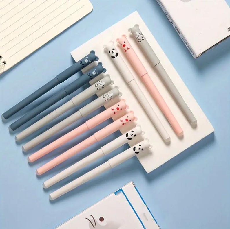 May include: A collection of colorful pens with animal-themed toppers. The pens are arranged on a white surface against a light blue background. The pens feature bear, panda, and cat designs in various colors, including blue, pink, gray, and white.