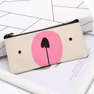 May include: A rectangular canvas pencil case with a black zipper. The case features a light beige background with a pink circle and black details resembling a bear's face. Two black dots are on either side of the pink circle.