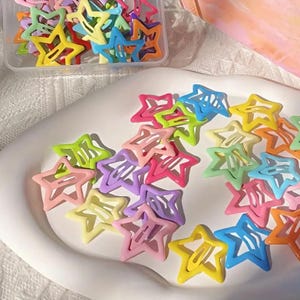 May include: A collection of colorful star-shaped hair clips in various shades, including pink, yellow, blue, and green. Some clips are scattered on a white surface, while others are in a clear plastic container. These clips are a fun accessory.