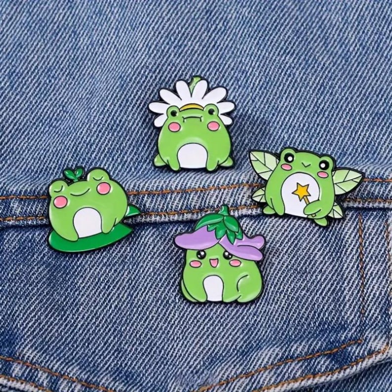 May include: Four enamel pins featuring cartoon frogs in various designs. One frog wears a daisy crown, another sits on a lily pad, a third has fairy wings and a star wand, and the last wears a purple flower hat. The pins are green, white, and pink.