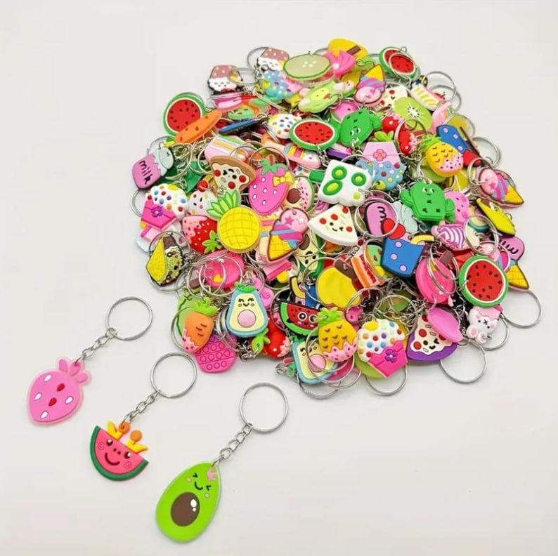 May include: A collection of colorful fruit-themed keychains, including strawberries, watermelons, avocados, and pineapples. Each keychain has a silver metal ring. The keychains are made of a soft, rubbery material.