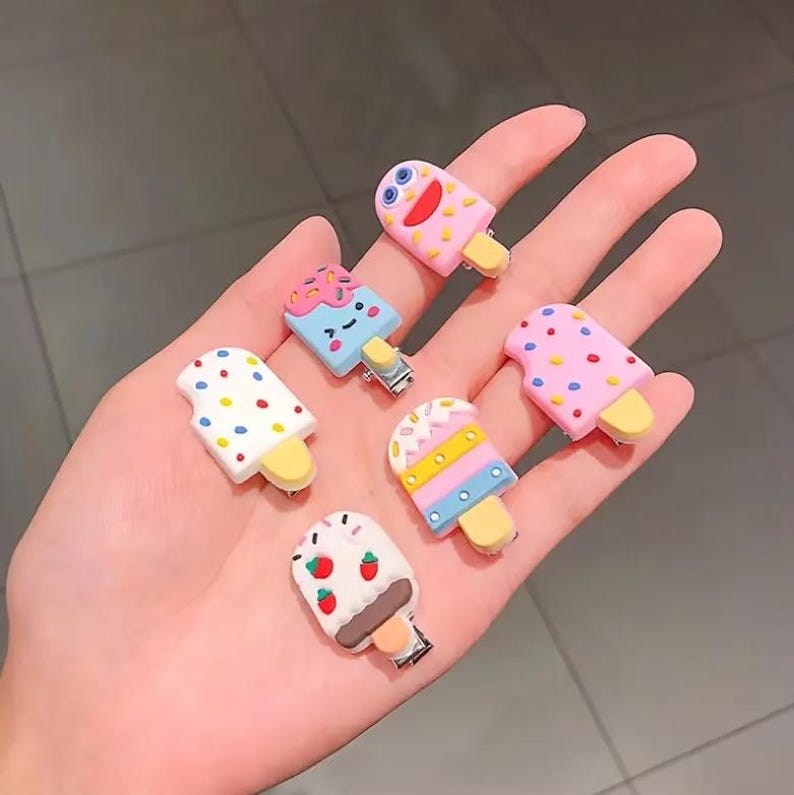May include: A collection of six colorful popsicle-shaped hair clips. Each clip features a different design, including sprinkles, dots, and fruit, with a yellow stick and a metal clip. The clips are in various shades of pink, blue, white, and yellow.