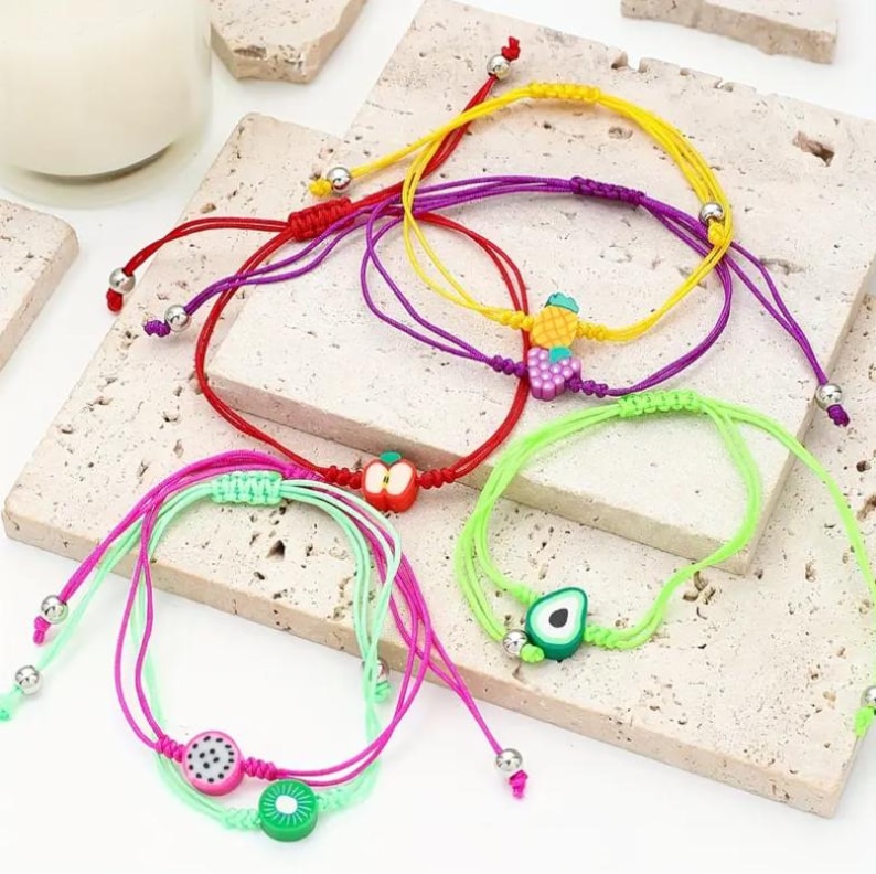 May include: A collection of colorful friendship bracelets with fruit charms. The bracelets feature red, yellow, purple, green, and pink cords. Each bracelet has a different fruit charm, including an apple, pineapple, grapes, avocado, dragon fruit, and kiwi.