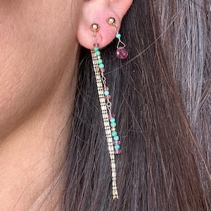 May include: A pair of gold earrings with a long chain featuring turquoise and pink beads. The earrings have a small pink gemstone dangling from the bottom.