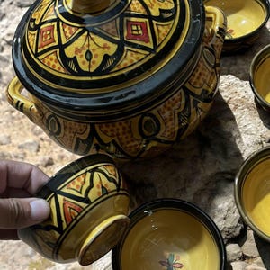 May include: A yellow and black ceramic tagine pot with matching bowls. The pot and bowls feature intricate geometric patterns in yellow, black, and red. The tagine has a lid and handles.