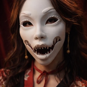 Puede incluir: Una máscara blanca con una boca con dientes afilados y aberturas para los ojos. La máscara se usa con una gargantilla roja y una blusa con estampado floral. La persona tiene cabello castaño largo y aretes dorados.