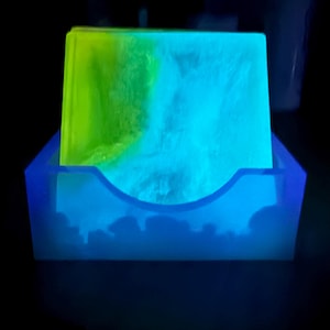 May include: A glowing blue resin coaster holder with a stack of coasters. The coasters are a mix of neon green and blue, creating a vibrant, luminous effect. The holder has a unique, curved design.