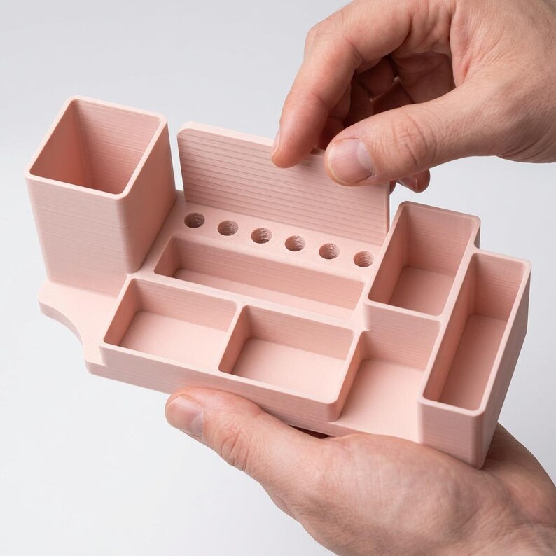 May include: A light pink desk organizer with multiple compartments, including a pen holder, small trays, and a hinged lid. The organizer is held by hands, showcasing its design and functionality. The item is made of a solid material.