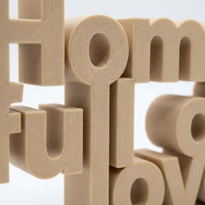 K&ouml;nnte beinhalten: Ein beiges, 3D-gedrucktes Dekoschild mit dem Wort "HOME". Die Buchstaben sind blockartig und gestapelt, wodurch ein geschichteter Effekt entsteht. Das Schild besteht aus einem festen, strukturierten Material und ist vor einem wei&szlig;en Hintergrund platziert.
