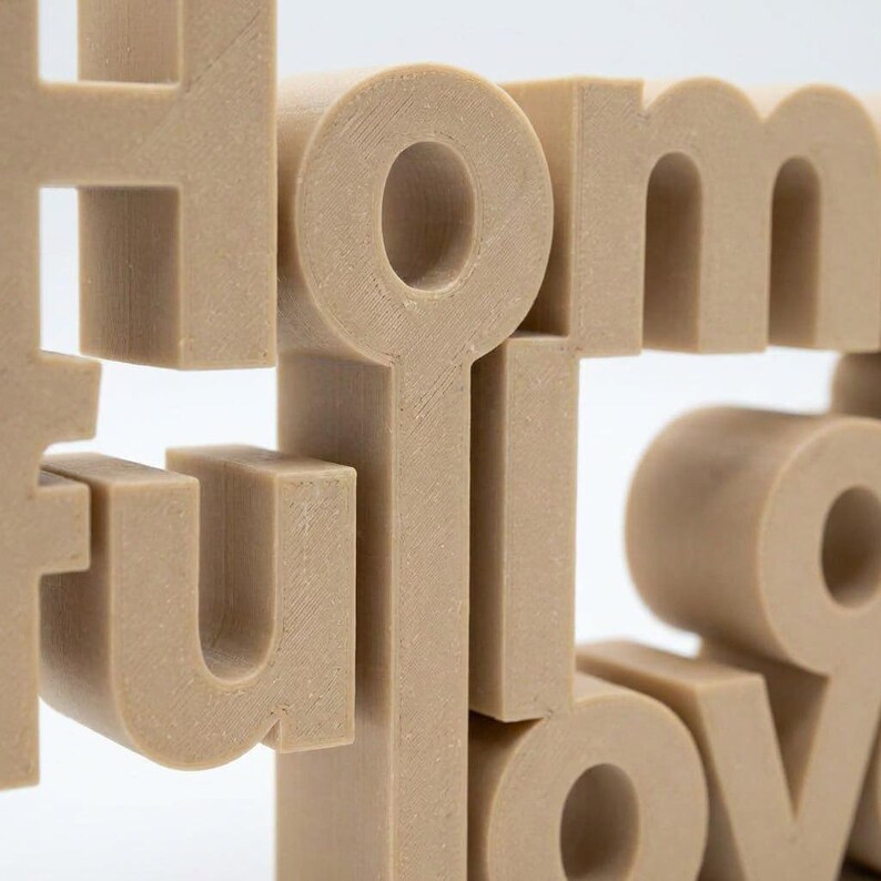K&ouml;nnte beinhalten: Ein beiges, 3D-gedrucktes Dekoschild mit dem Wort "HOME". Die Buchstaben sind blockartig und gestapelt, wodurch ein geschichteter Effekt entsteht. Das Schild besteht aus einem festen, strukturierten Material und ist vor einem wei&szlig;en Hintergrund platziert.