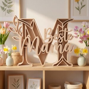 Op de afbeelding: Een lichtbruin houten decoratief bord in de vorm van de letter 'M' met de woorden 'Beste Mama' in cursief. Het bord staat op een houten plank met vazen ​​met bloemen op de achtergrond.