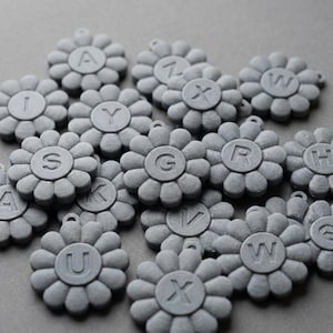 May include: A collection of gray, flower-shaped charms, each with a different letter of the alphabet in the center. The charms have a small loop at the top for attaching to a necklace or bracelet. The background is a solid gray.