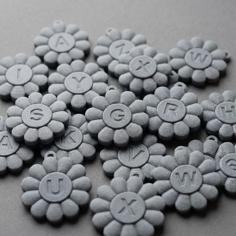May include: A collection of gray, flower-shaped charms, each with a different letter of the alphabet in the center. The charms have a small loop at the top for attaching to a necklace or bracelet. The background is a solid gray.