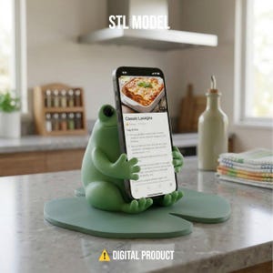 May include: A green frog phone holder on a lily pad base, holding a smartphone showing a lasagna recipe. The text "STL MODEL" and "DIGITAL PRODUCT" are displayed on the image.
