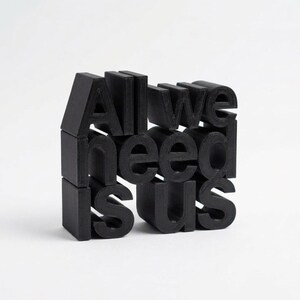 May include: Black 3D-printed sculpture spelling out "All we need is us" on a white background. The letters are blocky and evenly spaced, creating a modern, minimalist design. The sculpture is a decorative piece.