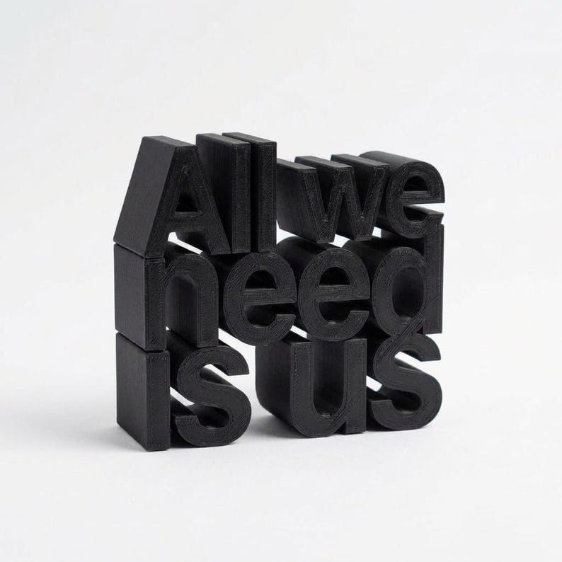 May include: Black 3D-printed sculpture spelling out "All we need is us" on a white background. The letters are blocky and evenly spaced, creating a modern, minimalist design. The sculpture is a decorative piece.