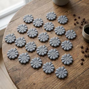 May include: A collection of gray, daisy-shaped silicone coasters, each with a different letter of the alphabet in the center. The coasters are arranged on a wooden table, with coffee beans and a mug in the background.
