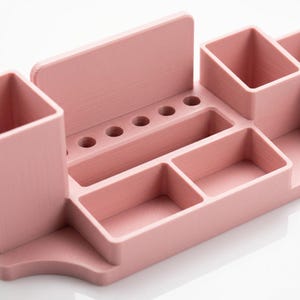 May include: A light pink desk organizer with multiple compartments. It features two square cup holders, a rectangular slot, and several small circular holes. The organizer is made of a solid material and has a smooth, matte finish.