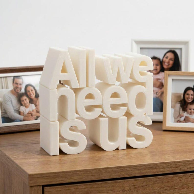 May include: A cream-colored, 3D sculpture with the phrase "All we need is us" displayed on a wooden surface. The letters are stacked and arranged to create a visually appealing design. Family photos are in the background.
