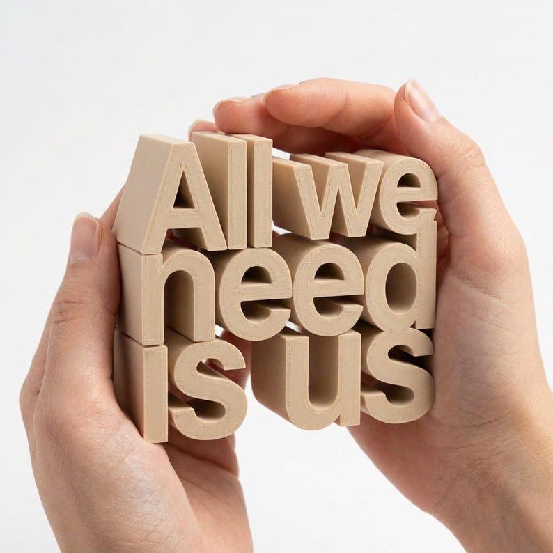 May include: A beige wooden block sculpture spelling out "All we need is us." The letters are in a sans-serif font and are held in two hands against a white background. The sculpture is a decorative item.