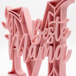 Op de afbeelding: Een lichtroze 3D-geprint decoratief stuk met de woorden "Beste Mama" in een cursief lettertype. Het ontwerp bevat de letter "M" met bloemaccenten. Het stuk is waarschijnlijk een Moederdag- of verjaardagscadeau.