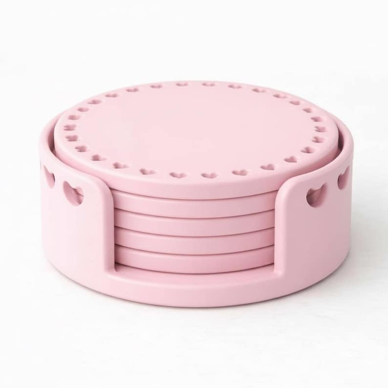 May include: A set of six pink coasters with a matching holder. Each coaster is round with a heart-shaped design around the edge. The holder has heart cutouts on the sides.
