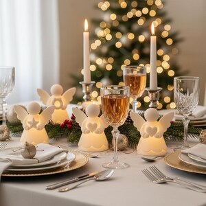 May include: A festive table setting with lit candles, angel figurines, and glasses of wine. The white angel figurines have glowing heart and star cutouts. The table is set with gold-rimmed plates, silverware, and a Christmas tree in the background.