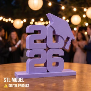 May include: A lavender 3D-printed graduation decoration featuring the year 2026 with a mortarboard hat. The base reads "STL MODEL" and "DIGITAL PRODUCT". The decoration is on a wooden table with a blurred background of people and lights.
