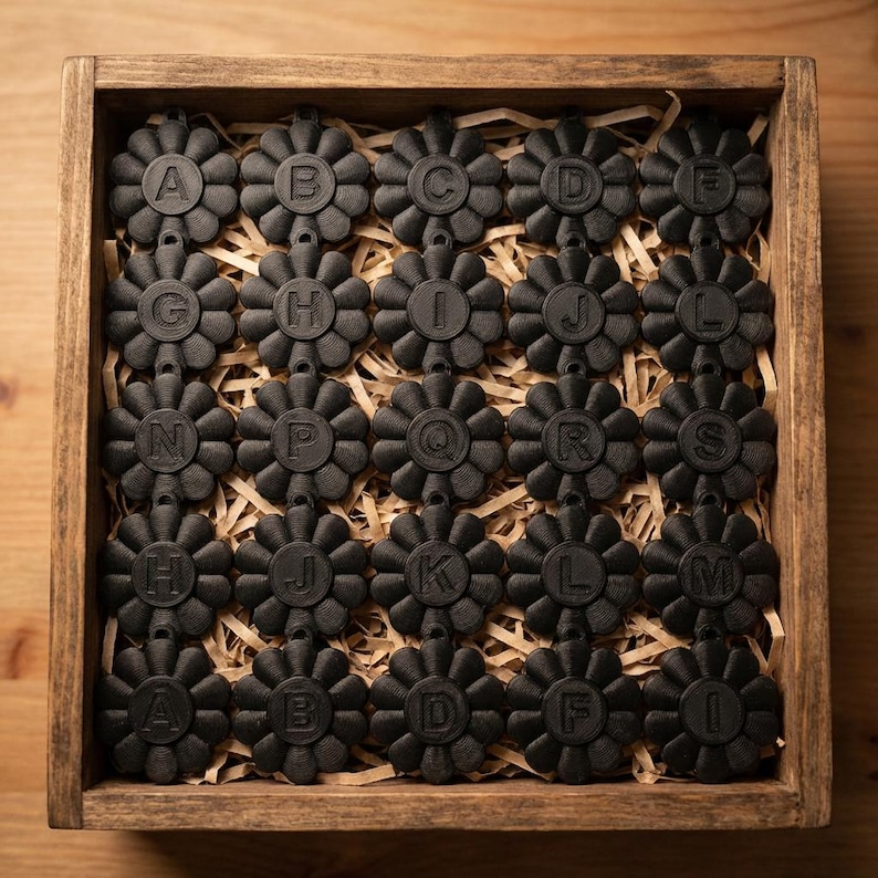 May include: A wooden box filled with black, flower-shaped alphabet charms. Each charm has a letter from the alphabet, from A to M, and is arranged on a bed of straw. The box is made of light brown wood.