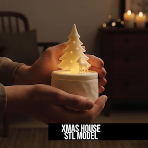 以下が含まれることがあります： 白のセラミッククリスマスハウス。点灯するクリスマスツリーと鹿のフィギュアが上にあります。ベースは円筒形です。「XMAS HOUSE STL MODEL」の文字が下にあります。