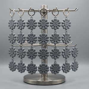 May include: A display of gray flower-shaped keychains, each with a different letter of the alphabet. The keychains are hanging on a silver metal display stand. The letters are in a sans-serif font. The background is a solid gray.