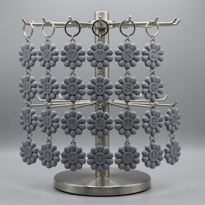 May include: A display of gray flower-shaped keychains, each with a different letter of the alphabet. The keychains are hanging on a silver metal display stand. The letters are in a sans-serif font. The background is a solid gray.
