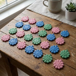 May include: A collection of colorful, flower-shaped alphabet charms in pink, blue, and green, arranged on a rustic wooden table. Each charm features a different letter of the alphabet. A mug and a small potted succulent are in the background.