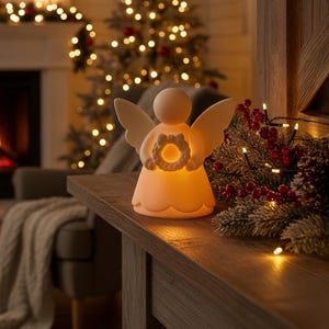May include: A glowing angel figurine with outstretched wings and a halo-like wreath. The off-white ceramic angel is illuminated from within, casting a warm glow. The angel is placed on a wooden surface, with Christmas lights and decorations in the background.