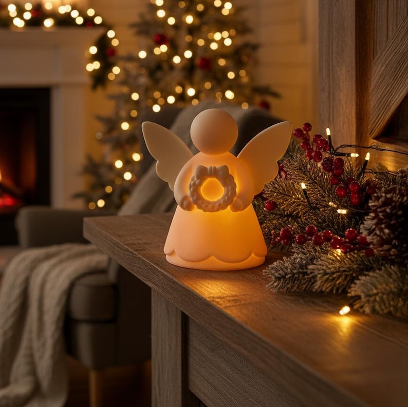 May include: A glowing angel figurine with outstretched wings and a halo-like wreath. The off-white ceramic angel is illuminated from within, casting a warm glow. The angel is placed on a wooden surface, with Christmas lights and decorations in the background.