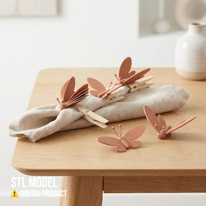 Op de afbeelding: Op een houten tafel ligt een beige linnen servet versierd met verschillende terracotta-kleurige vlindervormige servetringen. De servetringen zijn bevestigd aan houten wasknijpers. De tekst "STL MODEL" en "DIGITAL PRODUCT" is ook aanwezig.