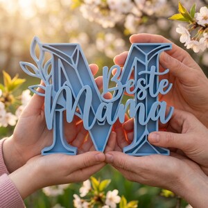Op de afbeelding: Een lichtblauw decoratief bord met de woorden "Beste Mama" in script, vastgehouden door twee paar handen. Het bord heeft de vorm van de letter "M" met bloemaccenten. De achtergrond is wazig met groen en zonlicht.