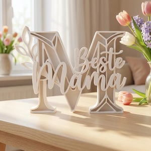 Op de afbeelding: Een cr&egrave;mekleurig houten decoratiestuk in de vorm van de letter "M" met de woorden "Beste Mama" in cursief. De letter is versierd met een bladontwerp. Het stuk staat op een houten tafel.