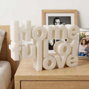 K&ouml;nnte beinhalten: Eine wei&szlig;e, dreidimensionale Skulptur mit der Aufschrift "Home full of love". Die Buchstaben sind in einer abgerundeten, serifenlosen Schriftart gehalten. Die Skulptur steht auf einem Nachttisch aus Holz, mit gerahmten Fotos im Hintergrund.